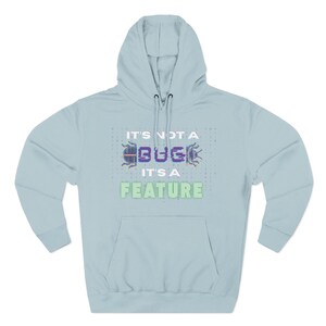 Peut inclure: Sweat à capuche bleu clair avec une poche avant et un cordon de serrage. Le devant du sweat à capuche affiche le texte "IT'S NOT A BUG IT'S A FEATURE" dans un design pixélisé. Le sweat à capuche a une capuche et des manches longues.