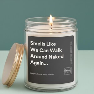 A candle in a clear or amber glass jar with a white label that reads “Smells Like We Can Walk Around Naked Again... Congratulations, empty nesters!!” placed on a simple, neutral background.