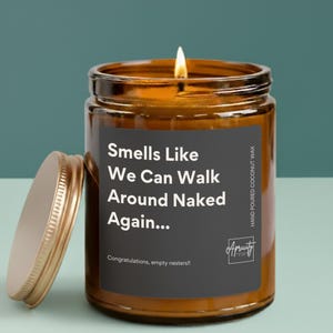 A candle in a clear or amber glass jar with a white label that reads “Smells Like We Can Walk Around Naked Again... Congratulations, empty nesters!!” placed on a simple, neutral background.