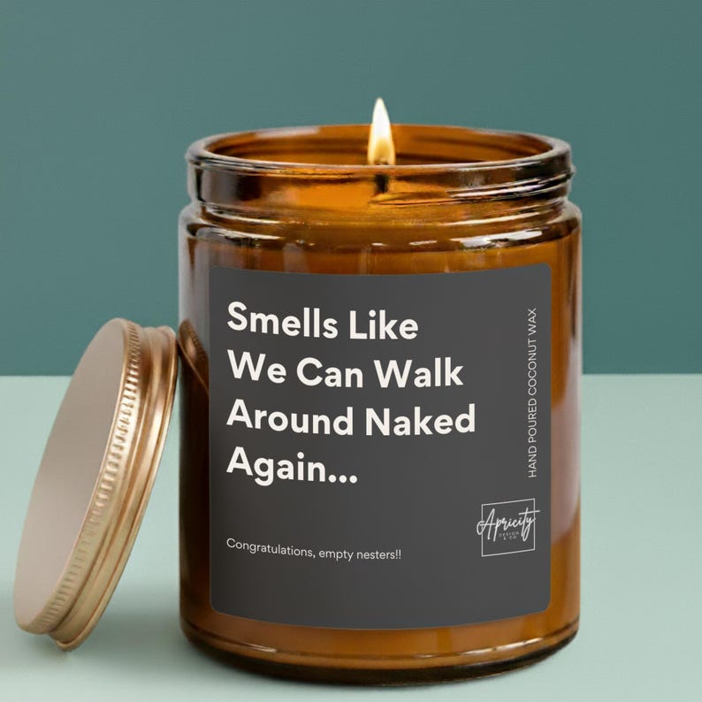 A candle in a clear or amber glass jar with a white label that reads “Smells Like We Can Walk Around Naked Again... Congratulations, empty nesters!!” placed on a simple, neutral background.