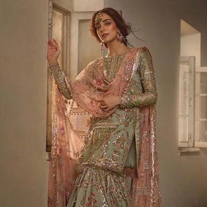 May include: An elegant, sage green ensemble with intricate gold embroidery and sequin embellishments. The outfit includes a long-sleeved top, a flared skirt, and a sheer, pink dupatta. The design features floral patterns and a matching headpiece.