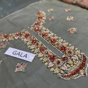 May include: Close-up of a sheer, gray fabric with intricate floral embroidery in shades of red, pink, and gold. The embroidery is concentrated around the neckline and along the edges. The word "GALA" is printed on a white label.