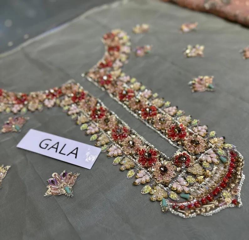 May include: Close-up of a sheer, gray fabric with intricate floral embroidery in shades of red, pink, and gold. The embroidery is concentrated around the neckline and along the edges. The word "GALA" is printed on a white label.