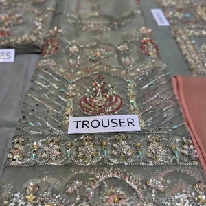 May include: Close-up of a gray embroidered fabric with intricate beadwork and embellishments. The fabric features a central design with red, gold, and green accents. The word "TROUSER" is printed on a white label.