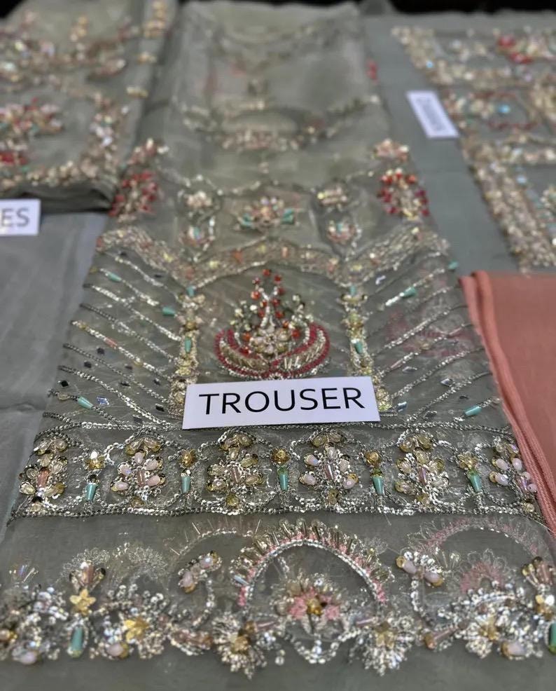 May include: Close-up of a gray embroidered fabric with intricate beadwork and embellishments. The fabric features a central design with red, gold, and green accents. The word "TROUSER" is printed on a white label.