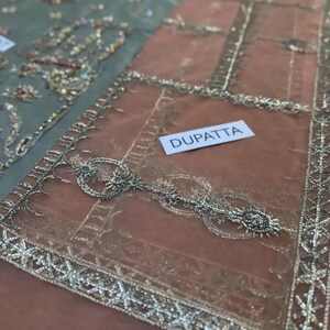 May include: Close-up of a sheer, peach-colored dupatta with intricate silver embroidery and embellishments. The word "DUPATTA" is printed on a white label. The dupatta has a detailed border design.