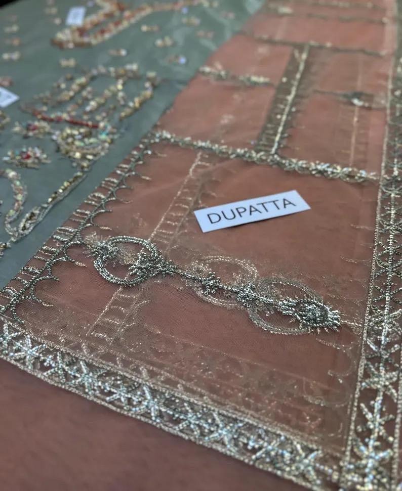 May include: Close-up of a sheer, peach-colored dupatta with intricate silver embroidery and embellishments. The word "DUPATTA" is printed on a white label. The dupatta has a detailed border design.