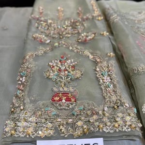 May include: A light green, sheer fabric with intricate embroidery. The embroidery features floral and geometric patterns in shades of gold, red, pink, and blue. The word "SLEEVES" is printed on a white label at the bottom.