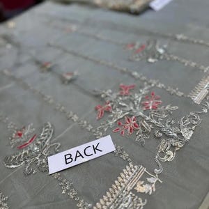 May include: Close-up of a sheer, gray fabric with intricate silver and red floral embroidery. The word "BACK" is printed on a white label. The fabric appears to be part of a garment, possibly a dress or top.