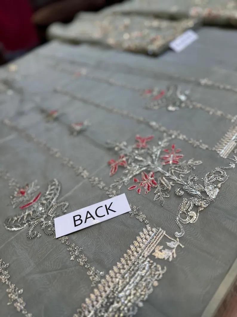 May include: Close-up of a sheer, gray fabric with intricate silver and red floral embroidery. The word "BACK" is printed on a white label. The fabric appears to be part of a garment, possibly a dress or top.