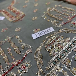 May include: Close-up of a gray fabric with intricate embroidery. The embroidery features gold, red, and multicolored beads and sequins, creating ornate floral and geometric patterns. A white tag reads "FRONT".