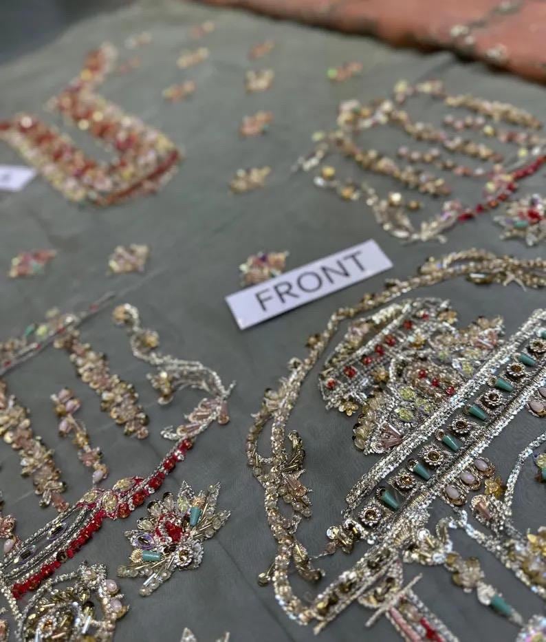 May include: Close-up of a gray fabric with intricate embroidery. The embroidery features gold, red, and multicolored beads and sequins, creating ornate floral and geometric patterns. A white tag reads "FRONT".