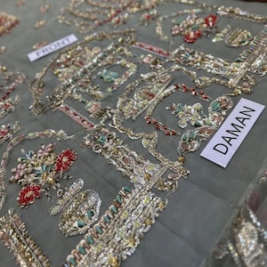 May include: Close-up of intricately embroidered fabric. The design features floral and geometric patterns in gold, red, and teal. The word "FRONT" is visible on a small white tag, and the word "DAMAN" is also visible on a white tag.