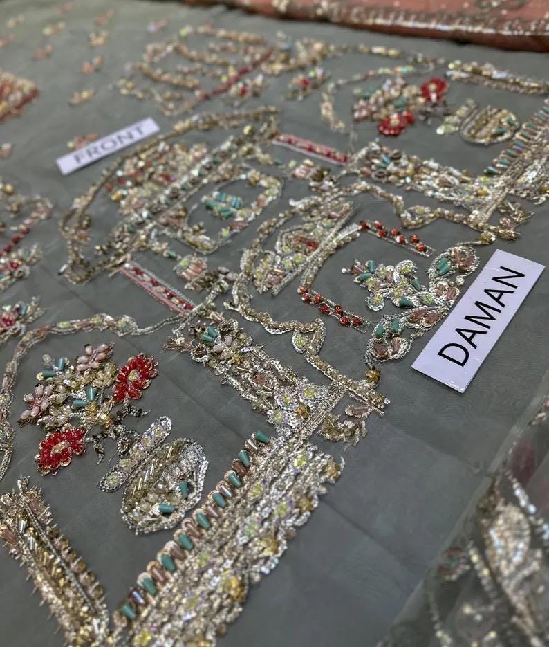 May include: Close-up of intricately embroidered fabric. The design features floral and geometric patterns in gold, red, and teal. The word "FRONT" is visible on a small white tag, and the word "DAMAN" is also visible on a white tag.