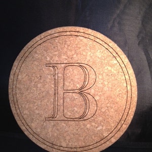 Puede incluir: Un posavasos redondo de corcho con un borde marrón oscuro y la letra "B" grabada en el centro en fuente serif.