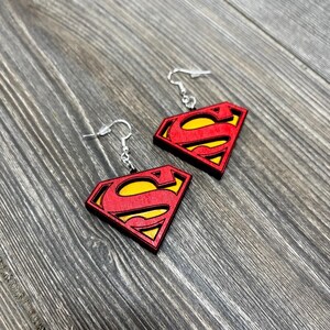 Puede incluir: Un par de pendientes de madera con el logotipo de Superman en rojo y amarillo. Los pendientes tienen ganchos plateados.