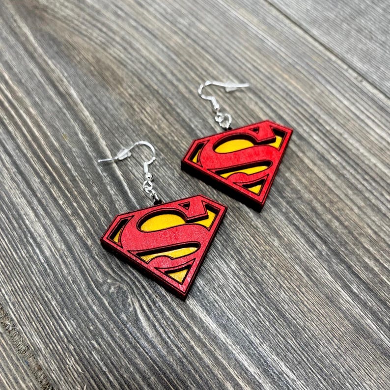 Puede incluir: Un par de pendientes de madera con el logotipo de Superman en rojo y amarillo. Los pendientes tienen ganchos plateados.