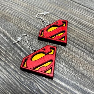 Puede incluir: Un par de pendientes de madera con el logotipo de Superman en rojo y amarillo. Los pendientes tienen un gancho plateado.