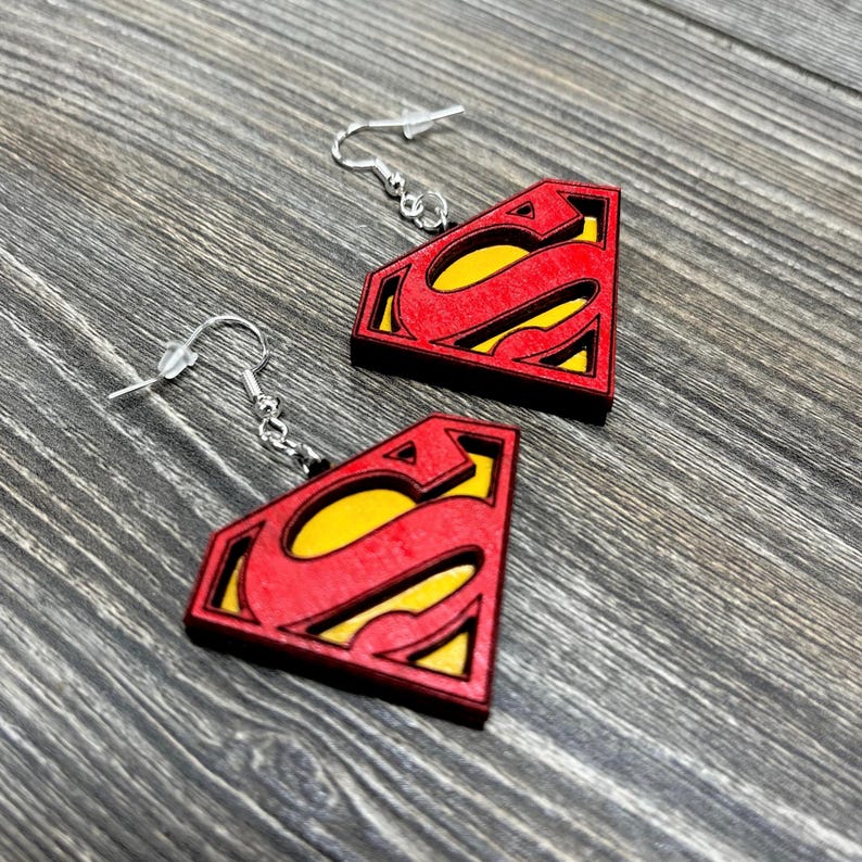 Puede incluir: Un par de pendientes de madera con el logotipo de Superman en rojo y amarillo. Los pendientes tienen un gancho plateado.
