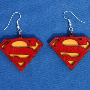 Puede incluir: Un par de pendientes de madera con el logotipo de Superman en rojo y amarillo. Los pendientes tienen un gancho plateado.