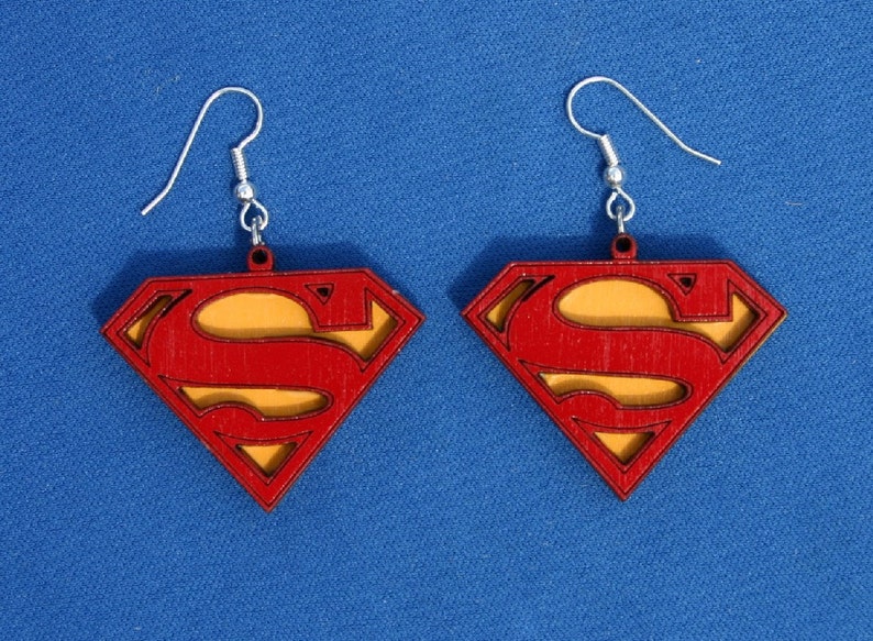 Puede incluir: Un par de pendientes de madera con el logotipo de Superman en rojo y amarillo. Los pendientes tienen un gancho plateado.