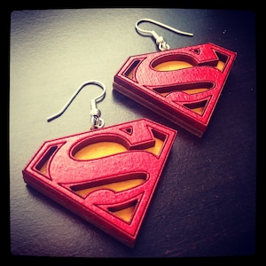 Puede incluir: Un par de pendientes de madera rojos y amarillos con forma del logotipo de Superman. Los pendientes est&aacute;n sobre una superficie marr&oacute;n oscura.