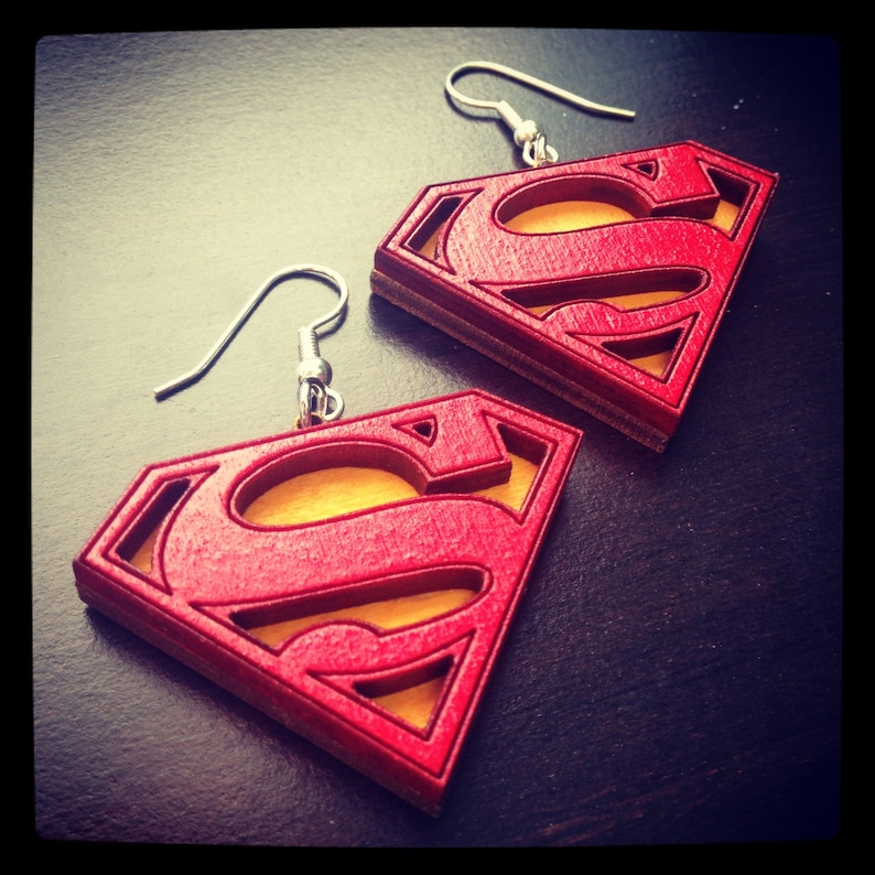 Puede incluir: Un par de pendientes de madera rojos y amarillos con forma del logotipo de Superman. Los pendientes est&aacute;n sobre una superficie marr&oacute;n oscura.