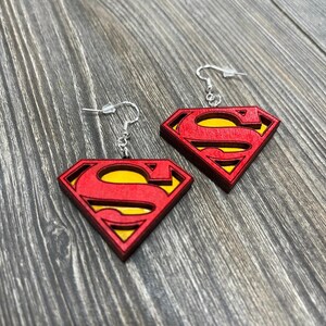 Puede incluir: Un par de pendientes de madera con el logotipo de Superman en rojo y amarillo. Los pendientes tienen un gancho plateado.