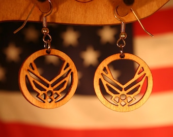 May include: A pair of wooden earrings with a cutout design of the United States Air Force emblem. The earrings are hanging from a wooden display stand against a red, white, and blue American flag background.