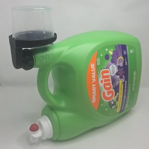 May include: A green laundry detergent bottle with a black measuring cup attached. The bottle has a white spout and a handle. The label reads "Gain" and "Smart Value". The measuring cup is clear plastic.
