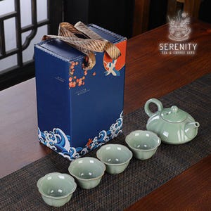 Op de afbeelding: Een theeset met een blauwe geschenkdoos met een kraanvogel- en golfontwerp. De set bevat een groene theepot en vier bijpassende kopjes. De doos heeft een gouden handvat. De tekst "SERENITY TEA & COFFEE SETS" is afgedrukt.