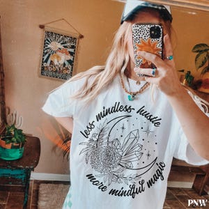 May include: White t-shirt with a black graphic design featuring a crescent moon, crystals, flowers, and the text "less mindless hustle more mindful magic." The shirt has short sleeves.