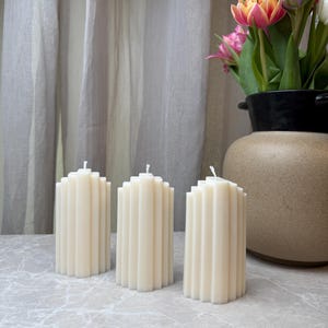 May include: Three off-white pillar candles with a vertical fluted, ribbed texture. Each candle features a cylindrical shape with a flat top and a single centered wick. The candles are arranged in a row, displaying a consistent wax finish.