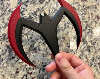 May include: A black and red bat-shaped object with a textured surface. The object has a central bat-like emblem and two curved, blade-like extensions with red edges. The item is held in a hand against a speckled background.