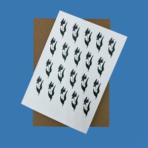 May include: A white card featuring a repeating pattern of dark blue flowers and green stems, placed on a brown card. The background is a solid blue. The floral design is simple and elegant, suitable for stationery or art prints.
