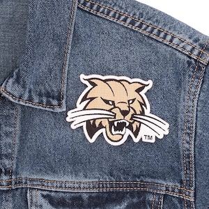 May include: A close-up of a blue denim jacket with a sewn-on patch. The patch features a tan and black wildcat design with the trademark symbol. The jacket has visible stitching.