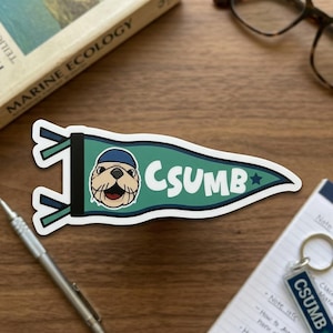 May include: A green pennant sticker with the text "CSUMB" in white. The pennant features a cartoon sea otter wearing a blue hat. A blue star is on the right side of the pennant. The sticker is on a wooden surface.