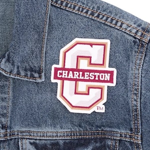 May include: Close-up of a blue denim jacket with a sewn-on patch. The patch features a white letter "C" with a maroon outline and the word "Charleston" in maroon. The jacket has visible stitching.