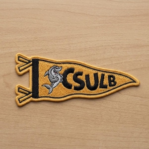 May include: Embroidered pennant patch in gold, black, and white. The patch features a smiling shark graphic and the letters "CSULB" in black. The patch has a yellow border and is likely for apparel or accessories.