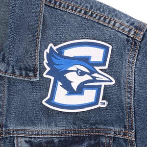 May include: Close-up of a denim jacket with a large patch featuring a blue jay bird logo. The patch is white, blue, and black, with the bird's head and the letter "G" incorporated into the design. The jacket has visible stitching.
