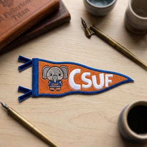 May include: Orange and blue pennant with the text "CSUF" in white. The pennant features an embroidered cartoon elephant wearing a CSUF uniform. The pennant is a decorative item.