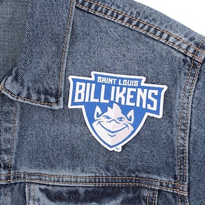 May include: Close-up of a blue denim jacket with a "Saint Louis Billikens" patch. The patch is white and blue, featuring the team name and a mascot graphic. The jacket has visible stitching.