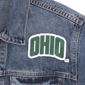 May include: Close-up of a blue denim jacket with the word "OHIO" embroidered in green and white on the front. The jacket has visible stitching and a classic design, suitable for casual wear.
