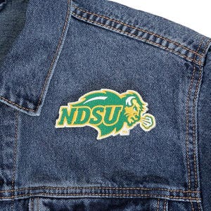 May include: Close-up of a blue denim jacket with a green and yellow NDSU logo patch on the chest. The patch features the letters "NDSU" and a bison graphic. The jacket has visible stitching.