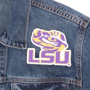May include: A close-up of a blue denim jacket with a patch featuring the LSU logo. The patch has a purple and yellow tiger graphic above the purple letters "LSU". The jacket has visible stitching.