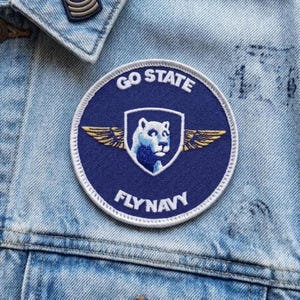 May include: A round, navy blue patch with the words "GO STATE" and "FLY NAVY" in white lettering. The patch features a white shield with a blue cougar head and gold wings. The patch is sewn onto a light blue denim jacket.