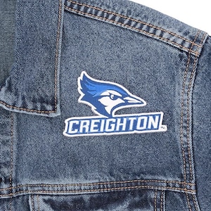 May include: Close-up of a denim jacket with a patch featuring a blue jay logo and the word "CREIGHTON" in white letters. The jacket has visible stitching and a classic denim texture.