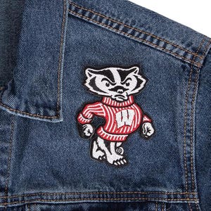 May include: A denim jacket with a patch featuring a cartoon badger in a red and white striped sweater. The badger has a stern expression and the letter "W" on its sweater. The patch is black, white, and red.