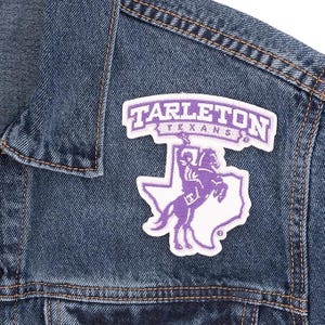 May include: Close-up of a blue denim jacket with a Tarleton Texans patch. The patch features the word "Tarleton" above "Texans" in purple and white. A purple and white graphic of a rider on a horse is below the text.