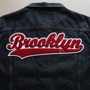 May include: A dark denim jacket with a large, embroidered "Brooklyn" patch in red and white. The patch is centered on the back of the jacket, showcasing a classic varsity style.